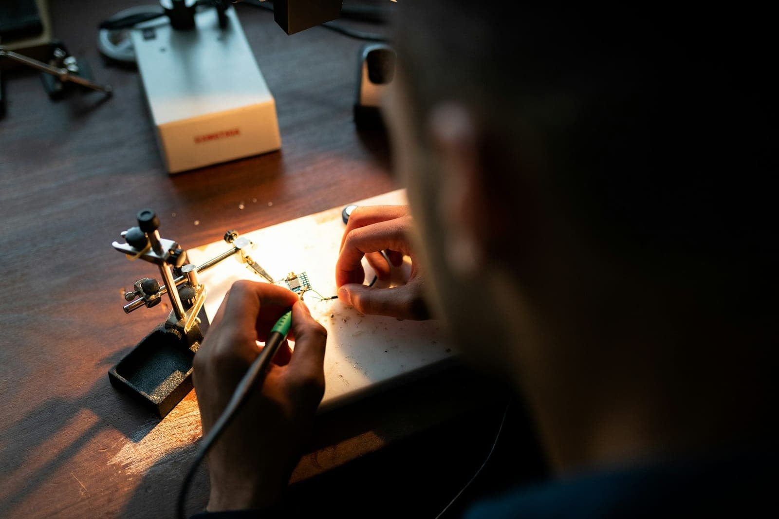Engineer seen over the shoulder soldering a small circuit board held in a helping-hands clamp under warm lamp light