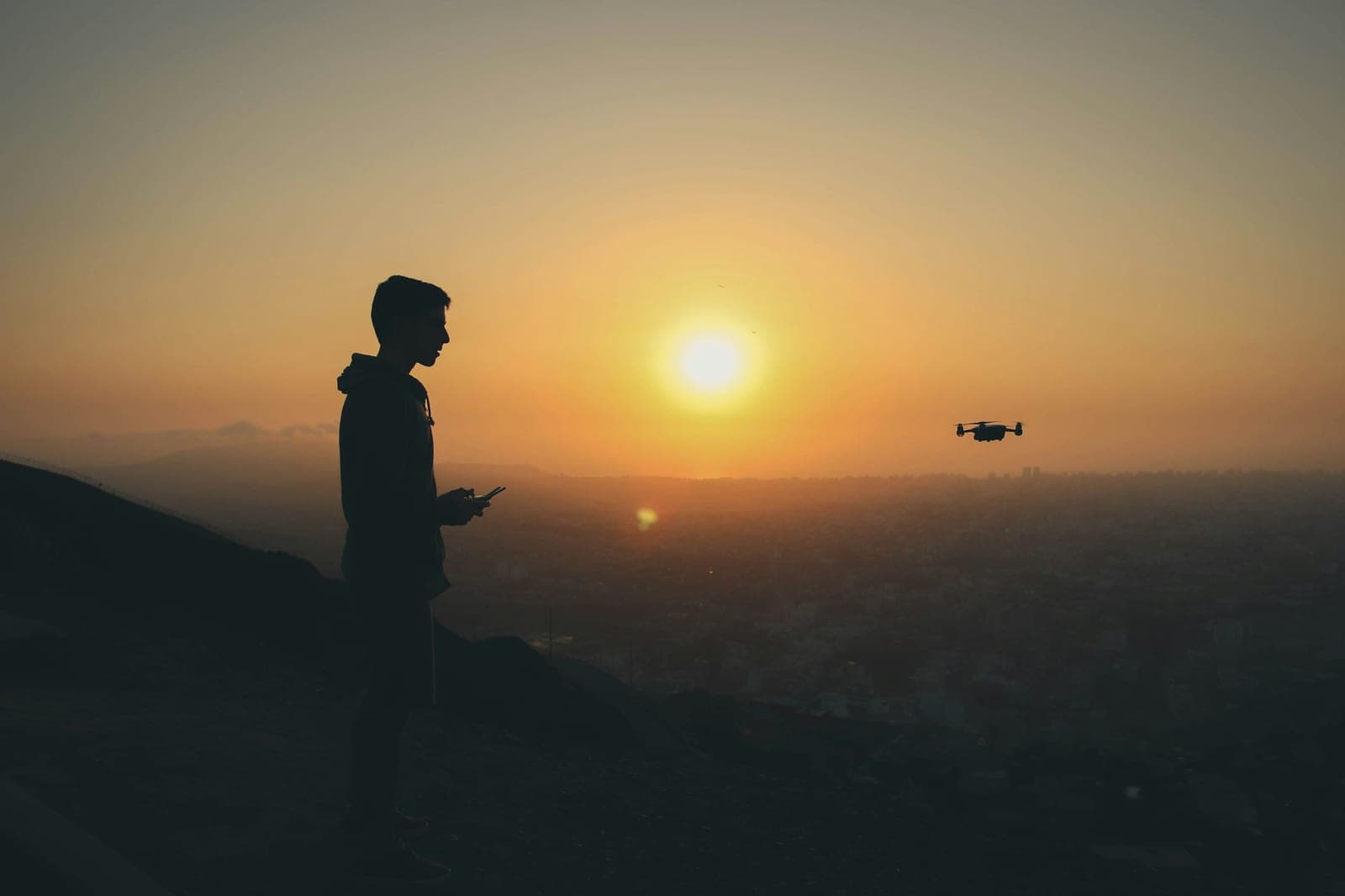 Silhouetted drone operator with a handheld controller on a hillside at sunset, a small quadcopter hovering nearby