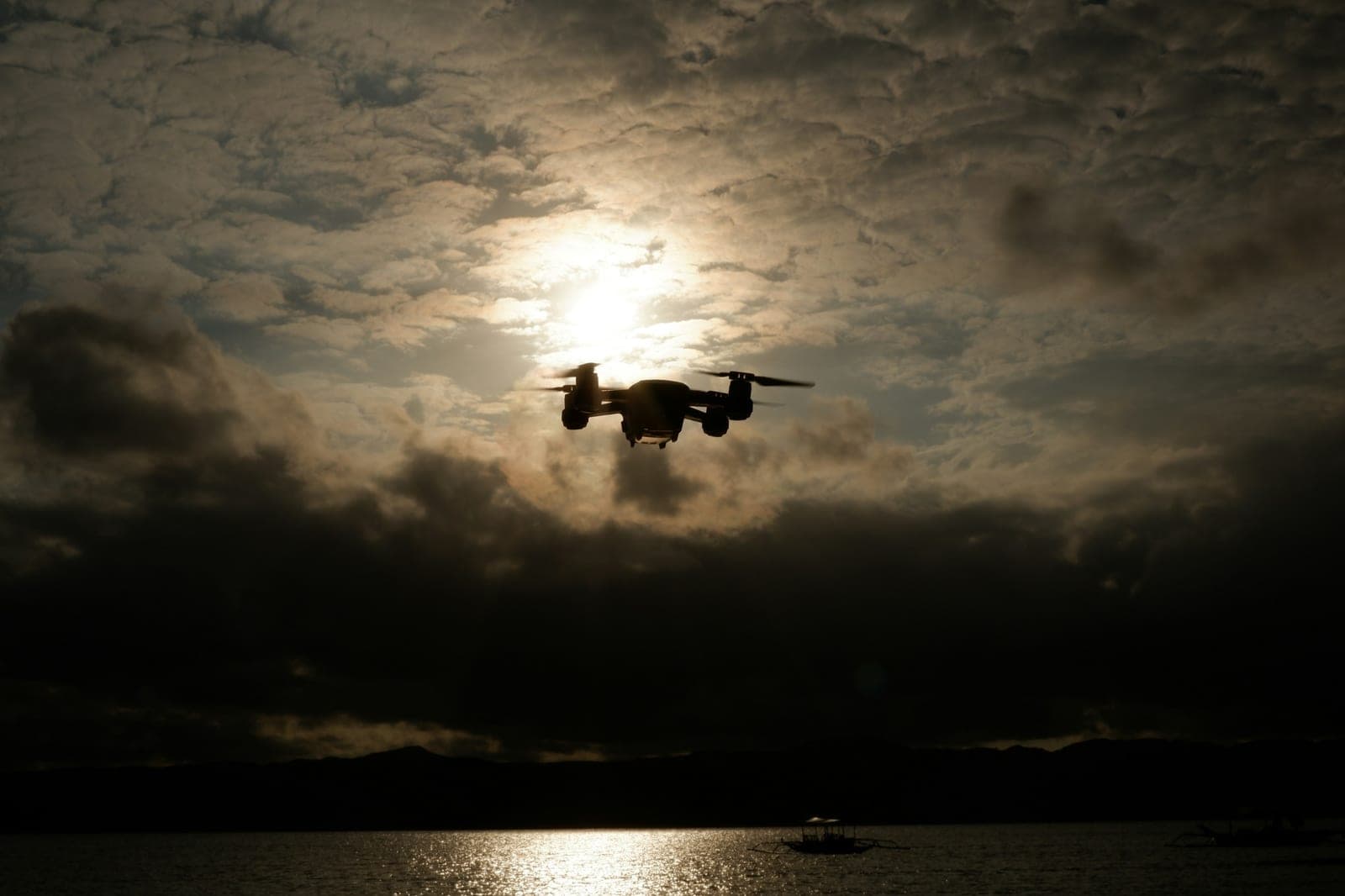 Small quadcopter drone silhouetted against a dark cloudy sky with the sun behind it, above the sea