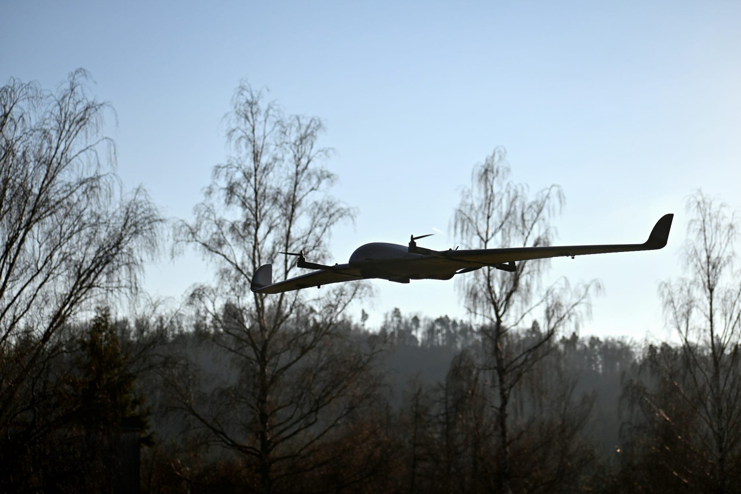 Fixed-wing VTOL drone flying low over a misty forest edge, backlit by a low winter sun