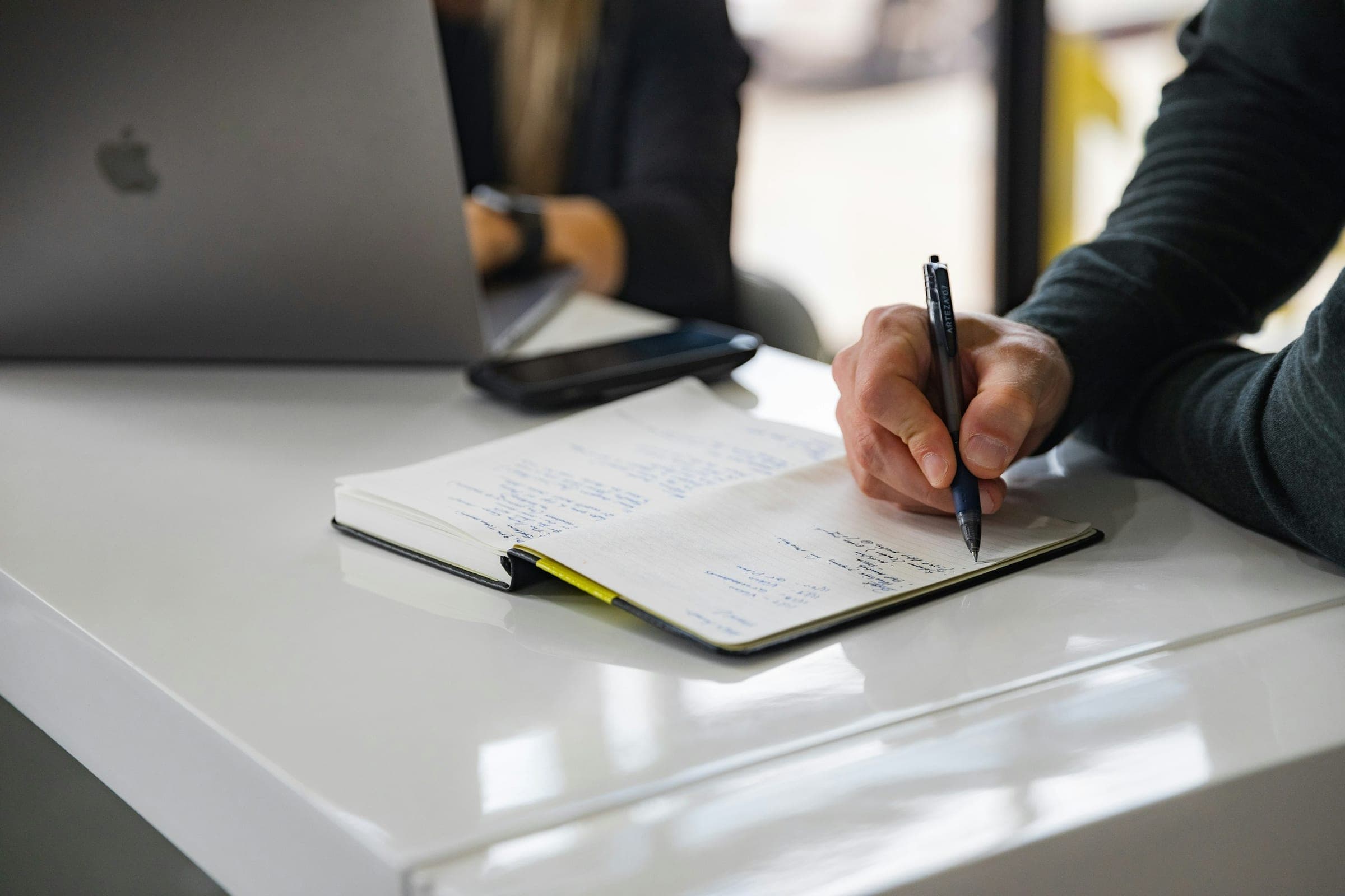 Hand writing notes in a notebook on a white table beside a laptop
