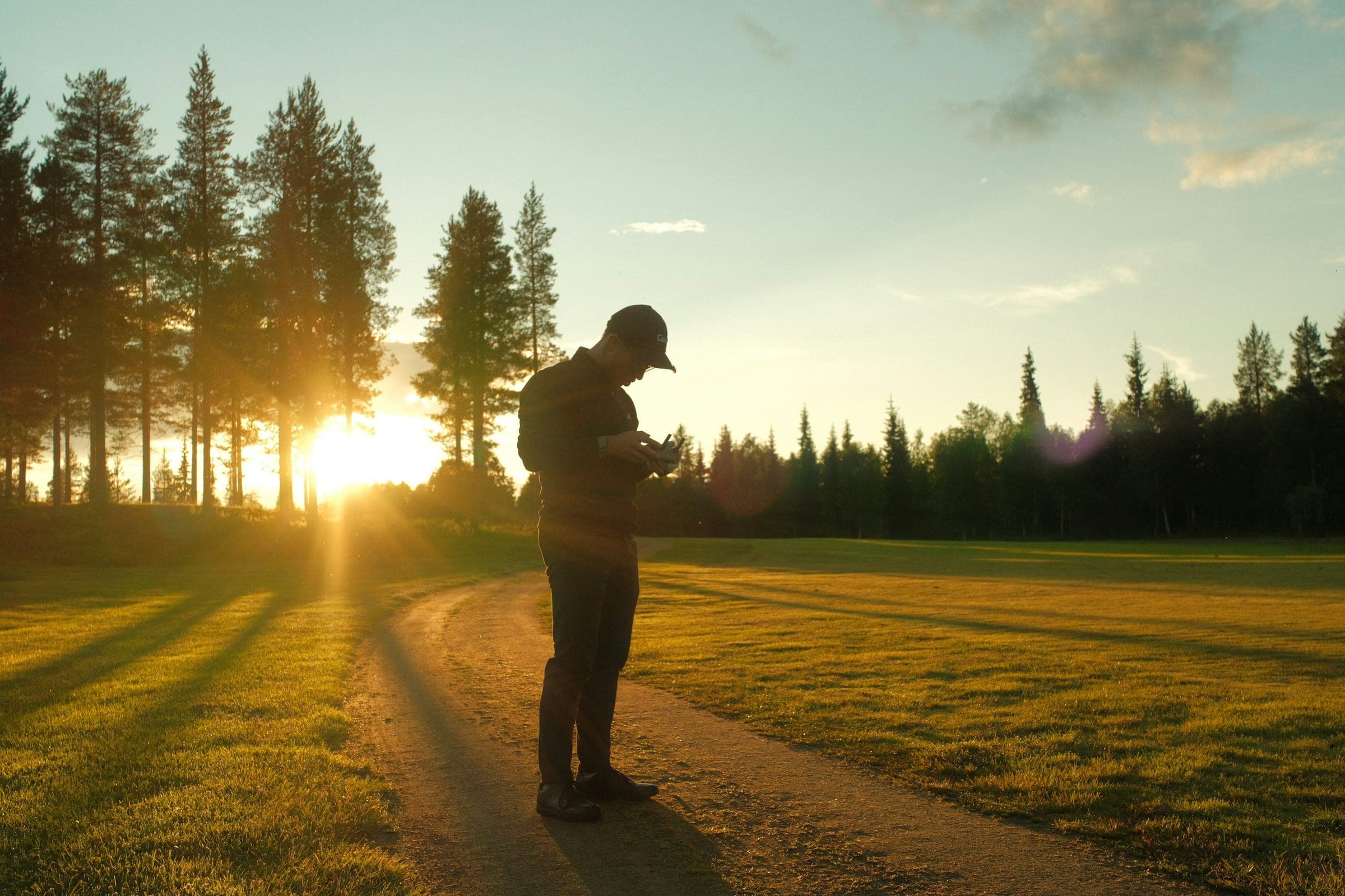 Person standing alone on a path through a field at sunset, holding a controller, pine trees behind