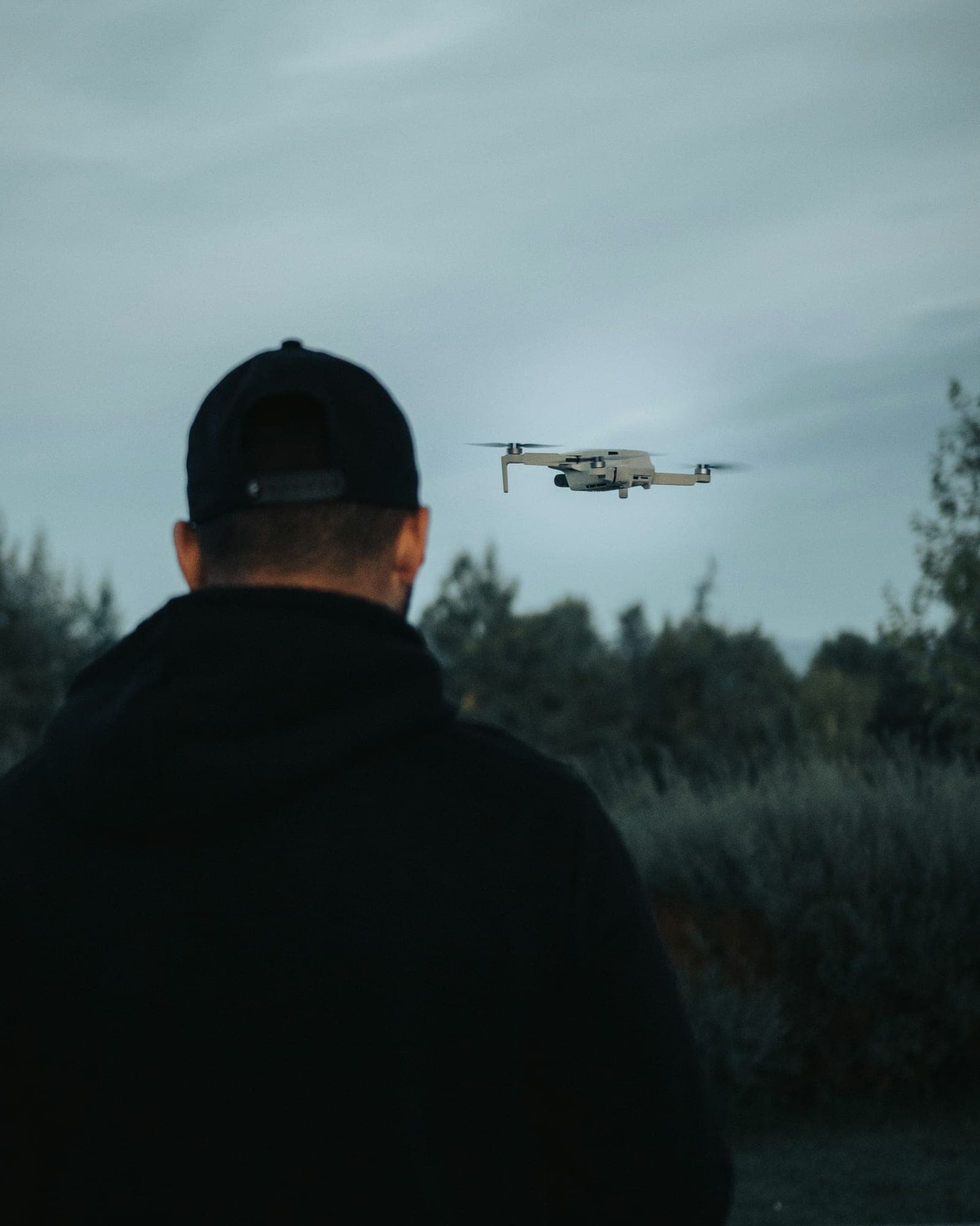 Operator in a dark jacket and cap, seen from behind, watching a small quadcopter hover at dusk