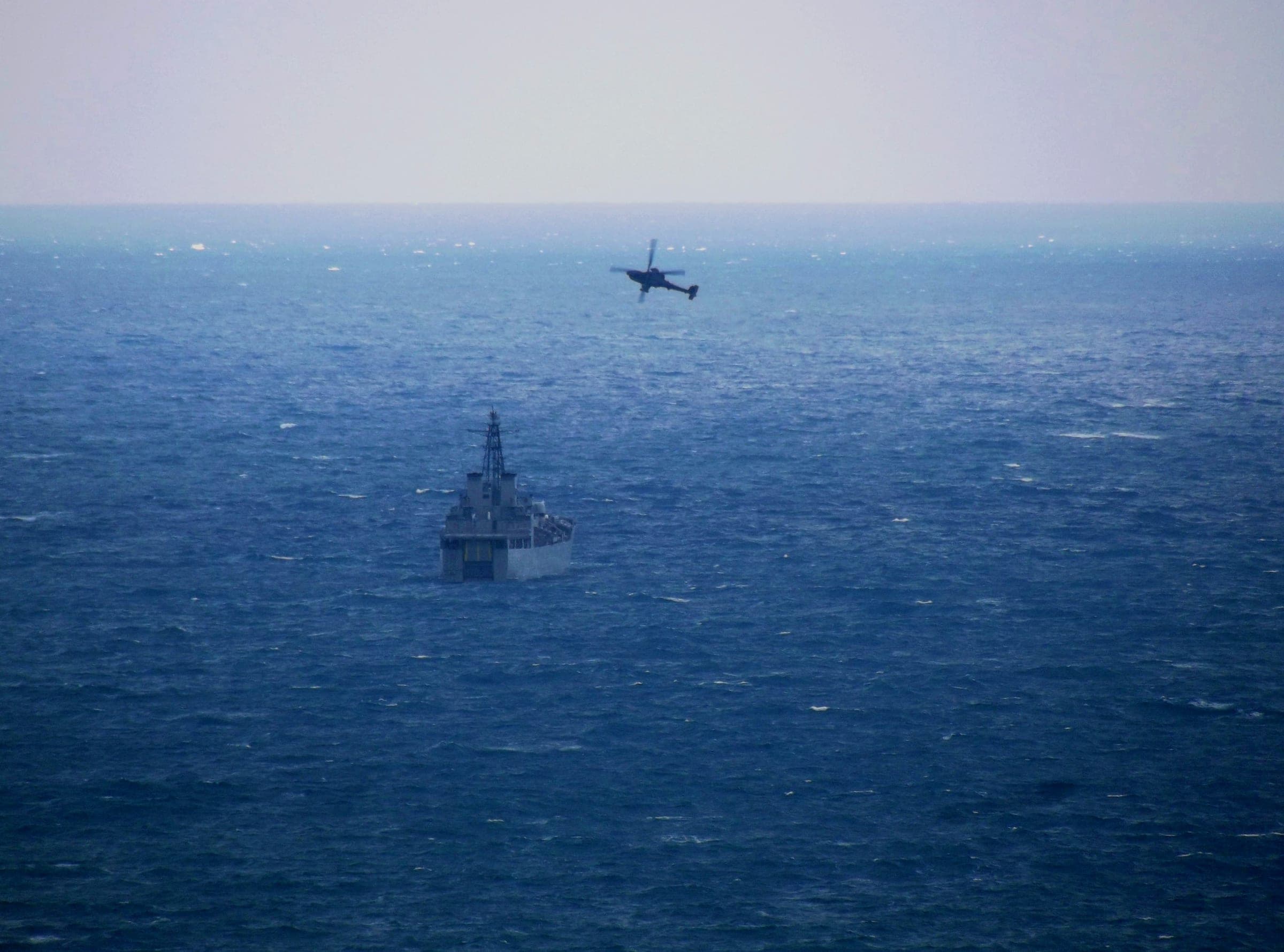 Gray naval vessel far out on open sea, a helicopter flying above it