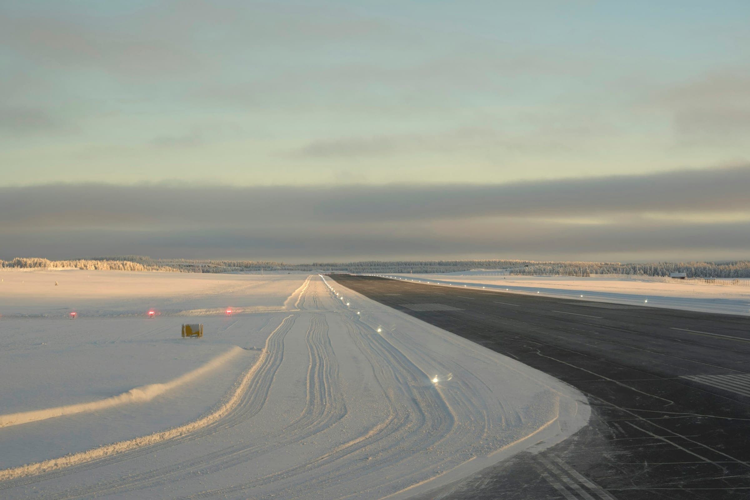 Snow-covered airfield at dawn, a cleared runway with edge lights stretching toward a frosted forest