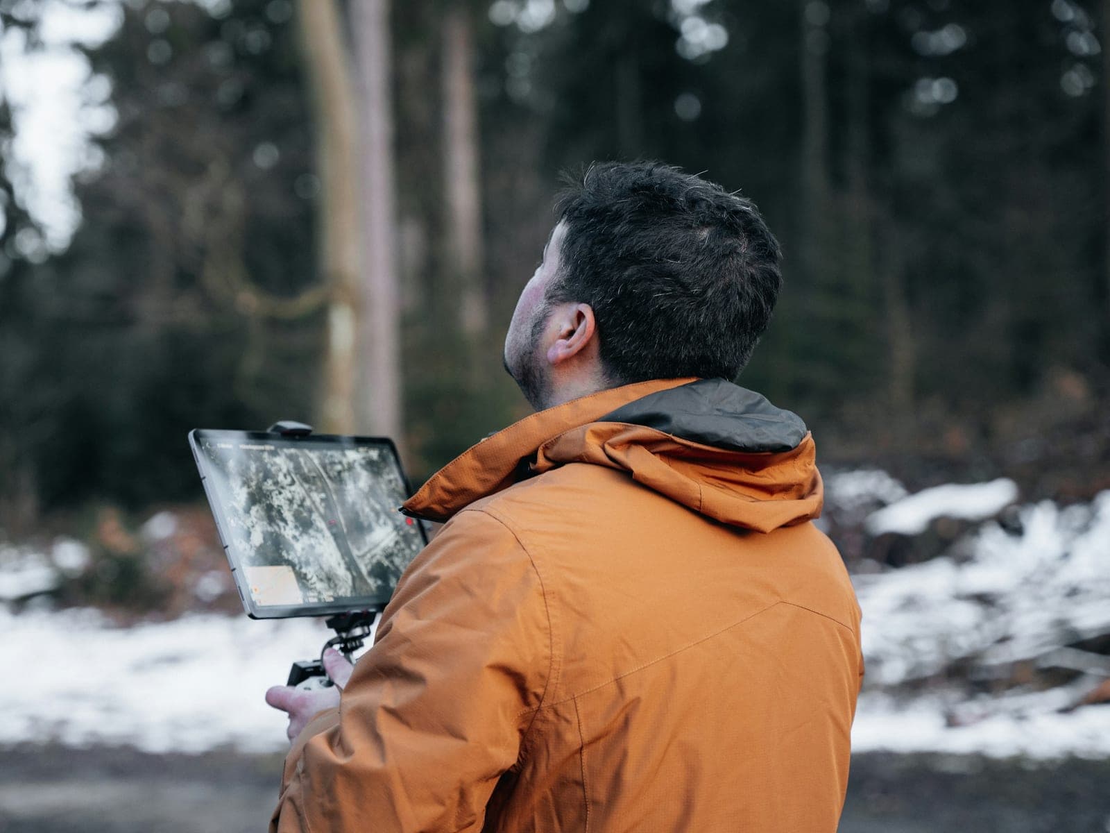Person in an orange jacket, seen from behind, holding a tablet on a controller showing aerial imagery in a snowy forest