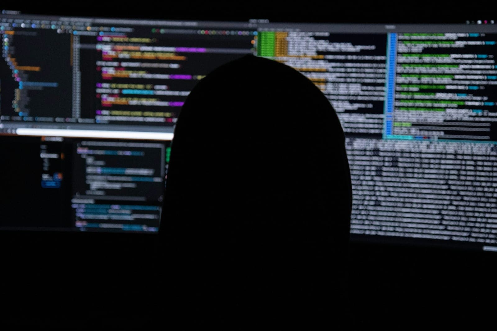 Silhouette of a developer in a dark room facing large monitors filled with out-of-focus code